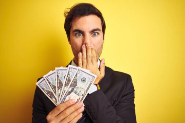 Takım elbiseli genç yakışıklı işadamı, dolarları izole edilmiş sarı arka planda tutuyor. Ağzı kapalı, utançtan ağzı açık, korkunun dışa vurumu, sessizlikten korkmak, gizli konsept.