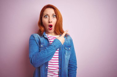 Güzel kızıl saçlı kadın kot gömlek ve çizgili tişört giyerek izole edilmiş pembe arkaplanı işaret ederek şaşırmış, ağzı açık şaşkın bir şekilde ifade ediyor..