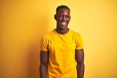 Gündelik tişört giyen genç Afro-Amerikalı adam izole edilmiş sarı arka planda dikiliyor kameraya seksi bir ifadeyle bakıyor, neşeli ve mutlu bir yüzle..