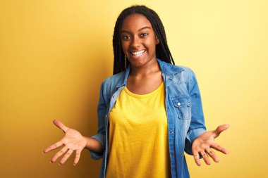 Genç Afro-Amerikalı kadın kot gömlek giyiyor, izole edilmiş sarı arka planda duruyor, neşeli gülümsüyor, kollarını açıp dostça karşılıyor, olumlu ve kendinden emin selamlar.