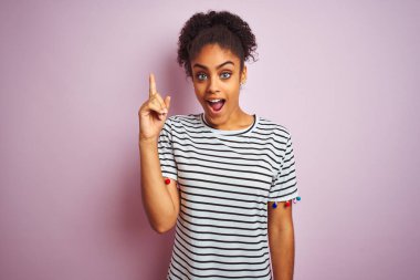 Siyah çizgili tişört giyen Afro-Amerikalı bir kadın izole edilmiş pembe arka planda durup başarılı bir fikri işaret ediyor. Heyecanlı ve mutlu. Bir numara..