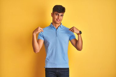 İzole edilmiş sarı arka planda duran mavi polo giyen genç Hintli adam yüzünde bir gülümsemeyle gururlu ve mutlu bir şekilde kendini işaret ediyor..