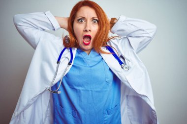 Genç, kızıl saçlı kadın steteskop kullanıyor, beyaz, soyutlanmış arka plan üzerinde. Deli ve elleri başında korkmuş, korkmuş ve ağzı açık şoktan şaşırmış.