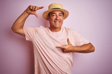 Yakışıklı, orta yaşlı, yaz şapkası takan, pembe arka planda duran, gülümseyen, elleri ve parmakları mutlu yüzlü bir adam. Yaratıcılık ve fotoğrafçılık kavramı.