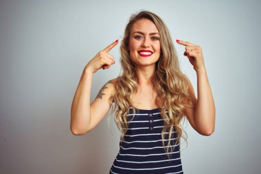 Çizgili tişört giyen genç ve güzel bir kadın beyaz izole edilmiş arka planda durup gülümsüyor iki eliyle başını işaret ediyor, harika bir fikir ya da düşünce, iyi bir hafıza.