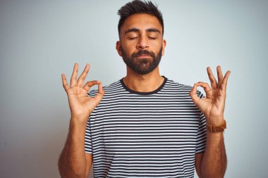 Siyah çizgili tişört giyen genç Hintli adam izole edilmiş beyaz arka planda dikiliyor rahatlıyor ve gözleri kapalı gülümsüyor parmaklarıyla meditasyon hareketi yapıyor. Yoga konsepti.