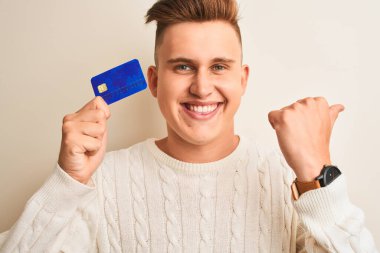Genç ve yakışıklı bir adam kredi kartını soyutlanmış beyaz arka planda tutuyor ve gülümseyen bir gülümsemeyle baş parmağıyla işaret ediyor.