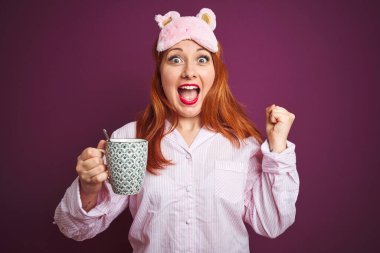 Genç kızıl saçlı kadın, pijama giymiş, mor izole edilmiş arka planda kahve içiyor gururla bağırıyor, zaferi kutluyor ve başarıyı kutluyor çok heyecanlı, coşkulu bir duygu.