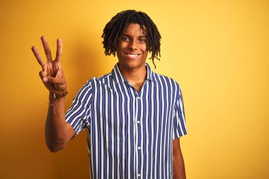 Rastgele çizgili tişörtler giyen Afro adam izole edilmiş sarı arka planda üç numarayı gösterirken kendinden emin ve mutlu bir şekilde gülümsüyor..