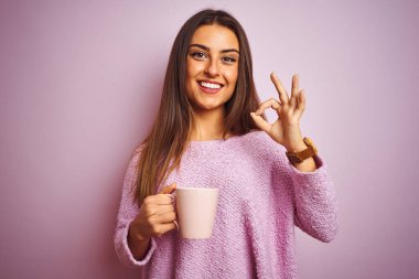 Bir fincan kahve içen genç ve güzel bir kadın izole edilmiş pembe arka planda duruyor. Tamam işareti yapıyor, mükemmel bir sembol.
