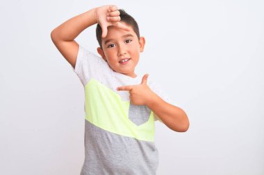 Güzel çocuk, izole edilmiş beyaz arka plan üzerinde sıradan bir tişört giyip gülümsüyor ve elleri ve parmakları mutlu yüzlü bir çerçeve yapıyor. Yaratıcılık ve fotoğrafçılık kavramı.