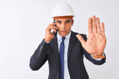 Miğfer takan genç mimar, akıllı telefonla konuşuyor. İzole edilmiş beyaz arka plan üzerinde. Açık elle dur işareti yapıyor.