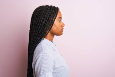 Genç Afro-Amerikan kadın çizgili gömlek giyiyor. İzole edilmiş pembe arka plan üzerinde duruyor. Yan yana bakıyor, rahat poz veriyor, kendine güvenen bir gülümsemesi var..