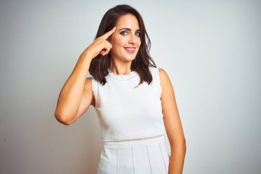 İzole edilmiş beyaz arka plan üzerinde duran elbiseler giyen genç ve güzel bir kadın gülümseyerek tek parmağıyla, harika bir fikirle ya da düşünceyle, iyi bir hafızayla başını işaret ediyor.