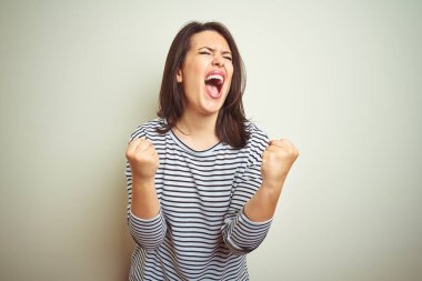 Genç ve güzel esmer kadın, beyaz izole edilmiş arka planda çizgili süveter giyiyor. Çok mutlu ve heyecanlı. Kollarını kaldırıp, gülümseyerek ve başarı çığlıkları atarak kazanan jestler yapıyor. Kutlama konsepti.