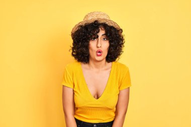 Kıvırcık saçlı, tişörtlü, şapkalı, izole edilmiş sarı arka planı olan genç bir Arap kadın korku, korku ve heyecan dolu bir yüz ifadesiyle dehşete düşmüş ve şok olmuştu..