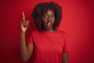 İzole edilmiş kırmızı arka planda duran ve başarılı bir fikri işaret eden tişört giyen Afrikalı genç bir kadın. Heyecanlı ve mutlu. Bir numara..