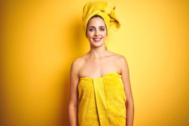 Güzel bir kadın vücuduna ve kafasına duş havlusu takmış mutlu ve serin bir gülümsemeyle izole edilmiş sarı bir arka plan. Şanslı kişi..