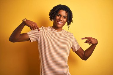 Sarışın, çizgili tişörtlü Afro adam izole edilmiş sarı arka planda duruyor. Yüzünde bir gülümsemeyle, gururlu ve mutlu bir şekilde kendini işaret ediyor..
