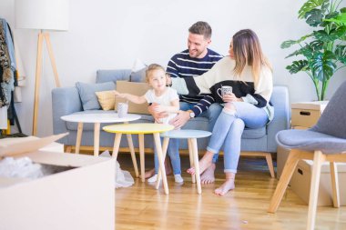 Güzel aile, anne-babalar kanepede kahve içme oturmuş karton kutular etrafında yeni evde oynayan çocuğuna bakıyor