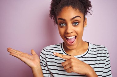 Siyah çizgili tişört giyen Afro-Amerikalı kadın izole edilmiş pembe arka planda el ve parmak göstererek mutlu bir şekilde duruyordu.