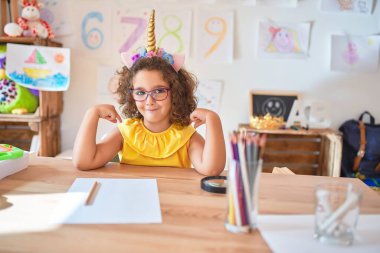 Gözlüklü ve tek boynuzlu at diademi takmış güzel bir çocuk anaokulunda masasına oturmuş, kendine güvenen, yüzünde bir gülümsemeyle, gururlu ve mutlu bir şekilde kendini işaret ediyor..
