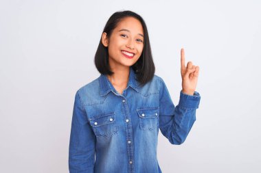 İzole edilmiş beyaz arka plan üzerinde duran kot gömlek giyen genç ve güzel Çinli kadın bir numaralı parmağını gösterirken kendinden emin ve mutlu bir şekilde gülümsüyor..
