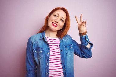Güzel kızıl saçlı kadın kot gömlek ve çizgili tişört giyerek izole edilmiş pembe arka planda gülümsüyor ve parmaklarını zafer işareti yaparken kameraya bakıyor. İki numara..