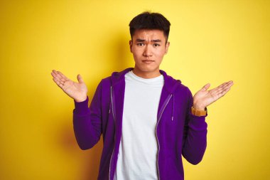 Mor kazak giyen Asyalı genç adam izole edilmiş sarı arka plan üzerinde duruyor. Kolları ve elleri havada, kafası karışmış bir ifade. Şüphe kavramı.