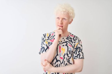 Renkli tişörtler giyen genç albino bir adam izole edilmiş beyaz arka planda duruyor ve elleri çenesinde soru sormayı düşünüyor. Düşünceli bir ifade. Düşünceli bir yüzle gülümsüyor. Şüphe kavramı.