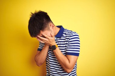 Asyalı, çizgili polo giyen genç bir adam izole edilmiş sarı arka planda dikiliyor ve ağlarken suratını ellerle kapatıyor. Depresyon kavramı.