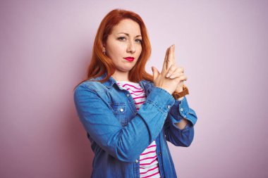 Güzel kızıl saçlı kadın kot gömlek ve çizgili tişört giyerek izole edilmiş pembe arka plan üzerinde sembolik bir silahla el hareketleriyle cinayet silahlarını tutuyor, kızgın surat ifadeleri sergiliyor.