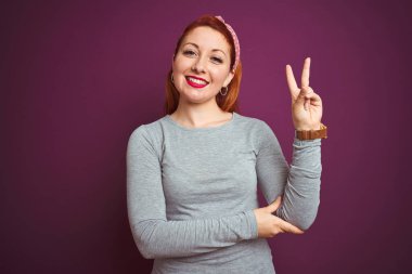 Gri tişört giyen güzel kızıl saçlı kadın ve izole edilmiş mor arka plan üzerinde tepe taklak duran, kameraya gülümseyen ve zafer işareti yapan bir kadın. İki numara..