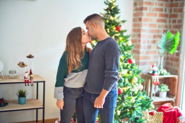 Mutlu ve kendinden emin gülümseyen genç bir çift. Evde Noel ağacının etrafında öpüşüp kucaklaşıyorlar.