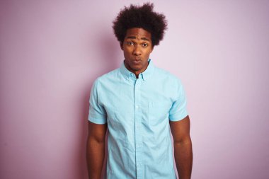 Afro saçlı, mavi tişörtlü genç Amerikalı adam izole edilmiş pembe arka plan üzerinde duruyor şüpheci ve gergin, sorun yüzünden üzgün. Negatif kişi.