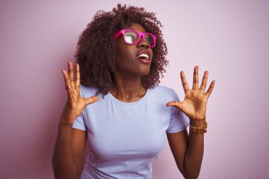 Genç Afrikalı Afro kadın pembe arka planda gözlük takıyor çılgın ve sinirli bir şekilde bağırıyor ve agresif bir ifade ve kolla bağırıyor. Hayal kırıklığı konsepti.