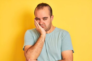 Yeşil tişört giyen genç adam izole edilmiş sarı arka planda duruyor. Kollarını kavuşturup depresyondan sıkılmış ve yorgun görünüyor..