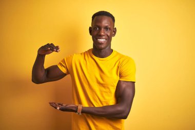 Sıradan tişörtler giyen genç Afro-Amerikan bir adam izole edilmiş sarı arka plan üzerinde duruyor ve elleri büyük ve büyük boy işareti, ölçü sembolü gösteriyor. Kameraya bakıp gülümsüyor. Ölçüm konsepti.