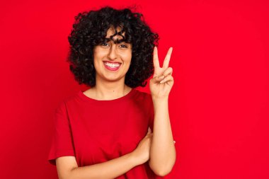 Kıvırcık saçlı genç Arap kadın izole edilmiş kırmızı arka plan üzerinde günlük tişört giyiyor ve kameraya gülümsüyor zafer işareti yapıyor. İki numara..