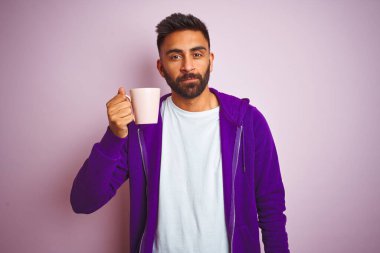 Mor kazak giyen Hintli bir adam izole edilmiş pembe arka planda bir fincan kahve içiyor. Zeki bir yüz ifadesiyle ciddi ciddi düşünüyor.