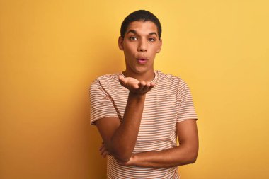 Çizgili tişört giyen genç yakışıklı Arap adam izole edilmiş sarı arka planda dikilip kameraya bakarak havada el ele öpüşerek güzel ve seksi görünmeye çalışıyor. Aşk ifadesi.
