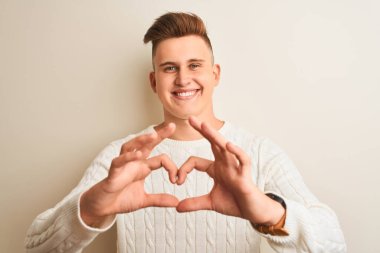 Kışlık süveter giyen yakışıklı genç adam izole edilmiş beyaz arka planda duruyor. Aşkla gülümsüyor. Elleriyle kalp sembolü ve şekli gösteriyor. Romantik konsept.