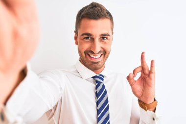 Genç yakışıklı iş adamı izole edilmiş arka planda akıllı telefon kullanarak selfie çekiyor. Tamam işareti yapıyor, mükemmel bir sembol.