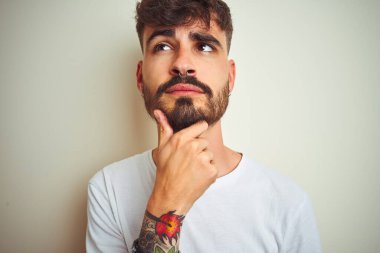 Dövmeli genç adam t-shirt giyen çene üzerinde el ile izole beyaz arka plan üzerinde duran soru, dalgın ifade düşünüyor. Düşünceli bir yüzle gülümsüyor. Şüphe kavramı.