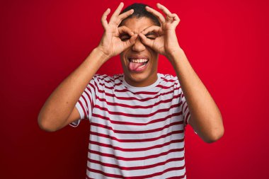 İzole edilmiş kırmızı arka plan üzerinde çizgili tişört giyen genç yakışıklı Arap adam dürbün gibi jest yapıyor, gözleri parmaklarından bakıyor. Çılgın ifade.