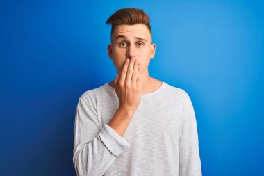İzole edilmiş mavi arka plan üzerinde duran beyaz tişörtlü genç yakışıklı bir adam. Ağzını kapayarak hata yaptığı için utancından, korkusunun ifadesinden, sessizlikten korkmasından, gizli konseptinden.