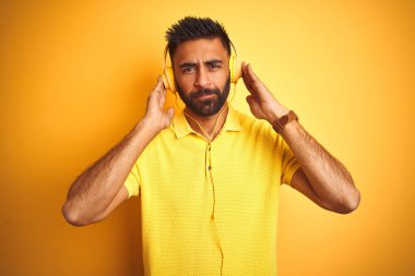 Arap Hindistanlı İspanyol adam müzik dinliyor. Kulaklık takıyor, sarı arka planda, kendine güvenen bir yüz ifadesi var. Ciddi düşünüyor.