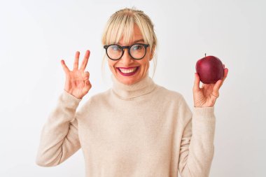 Elma tutan orta yaşlı bir kadın izole edilmiş beyaz arka planda duruyor. Tamam işareti yapıyor, mükemmel bir sembol.