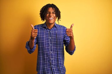 Rastgele tişörtlü Afro adam izole edilmiş sarı arka plan başarısı tabelasının üzerinde duruyor. Elleriyle olumlu jest yapıyor, gülümsüyor ve mutlu. Neşeli ifade ve kazanan jest.