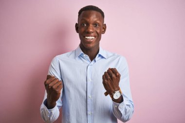 İzole edilmiş pembe arka plan üzerinde duran mavi şık gömlekli Afrikalı bir adam kollarını kaldırarak ve açık gözlerle başarıyı kutluyor ve şaşırıyor. Kazanan konsept.